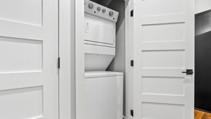 A laundry room with a stacked washer and dryer, white doors, and a wooden floor. - Custom Suite over Target - Downtown AU 209 (Auburn)