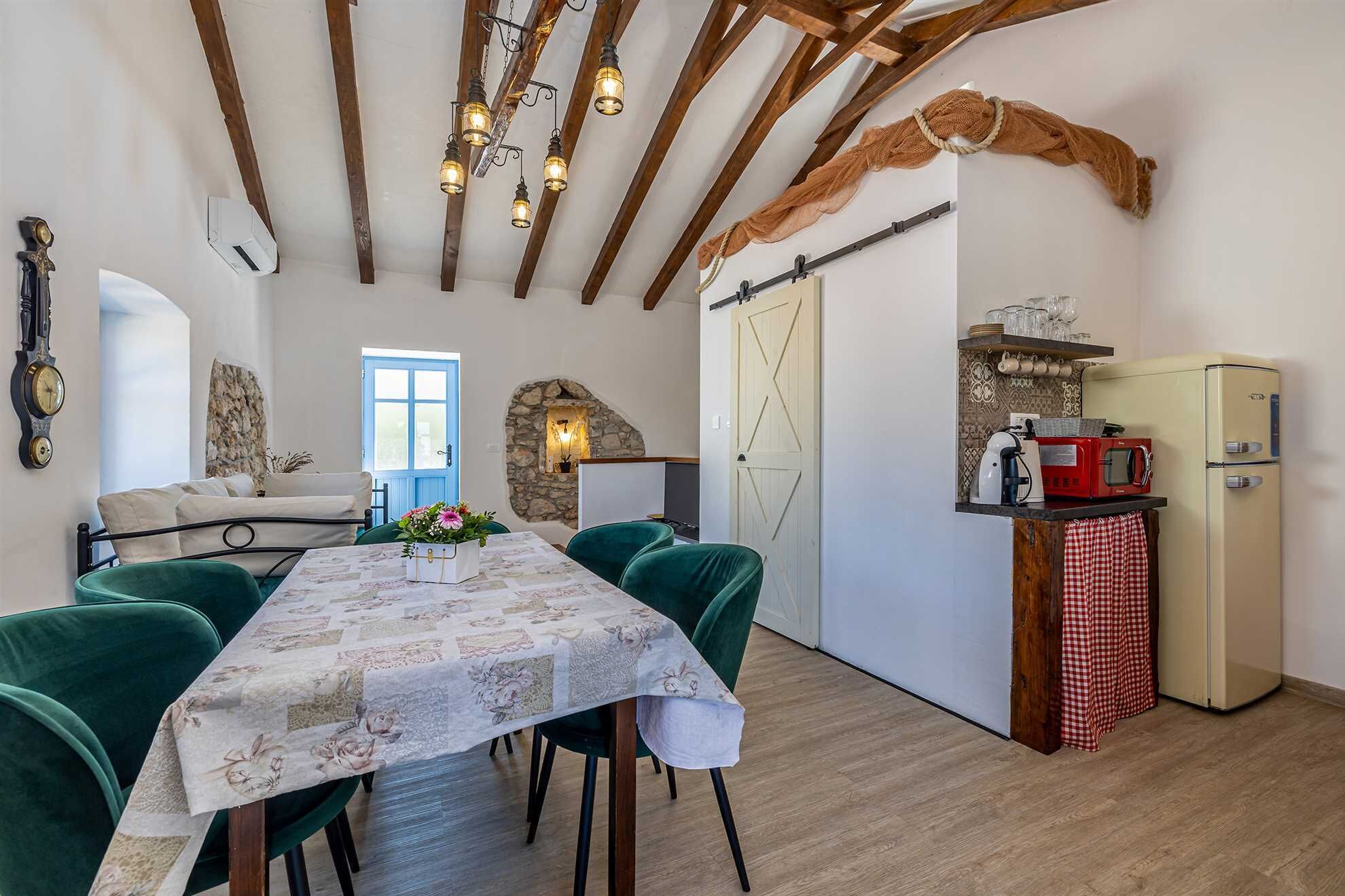 Un comedor con una mesa lista para cuatro, una cocina con un refrigerador antiguo y una pared de piedra con estufa empotrada.