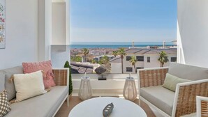 A modern living room with a large balcony, white sofas, wicker furniture, and a view of the sea and cityscape.