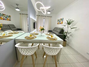 A modern dining area with a glass table, white chairs, and a ceiling fan. - Residencial Blaia  (Praia Grande)
