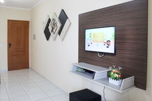 A modern living room with a flat-screen TV, a wooden accent wall, a white console table, and a door. - Residencial Blaia  (Praia Grande)