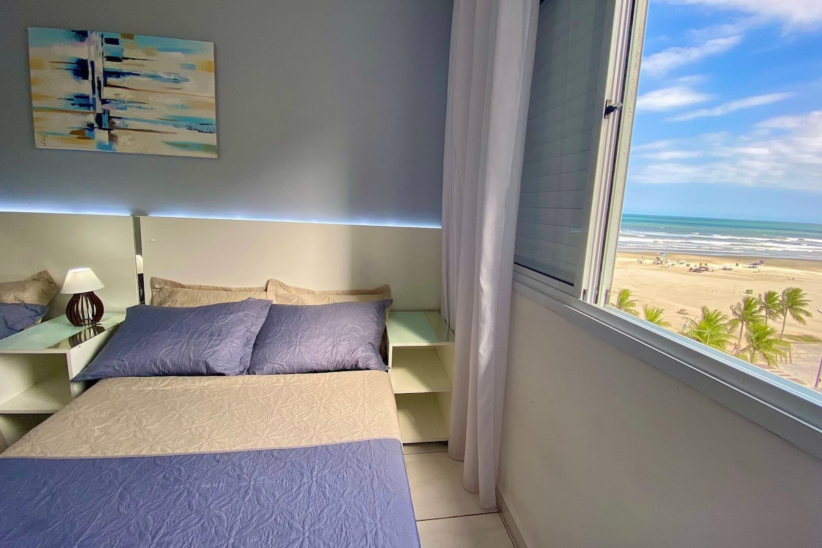 Quarto com cama, mesa de cabeceira, abajur, pintura e vista para a praia.