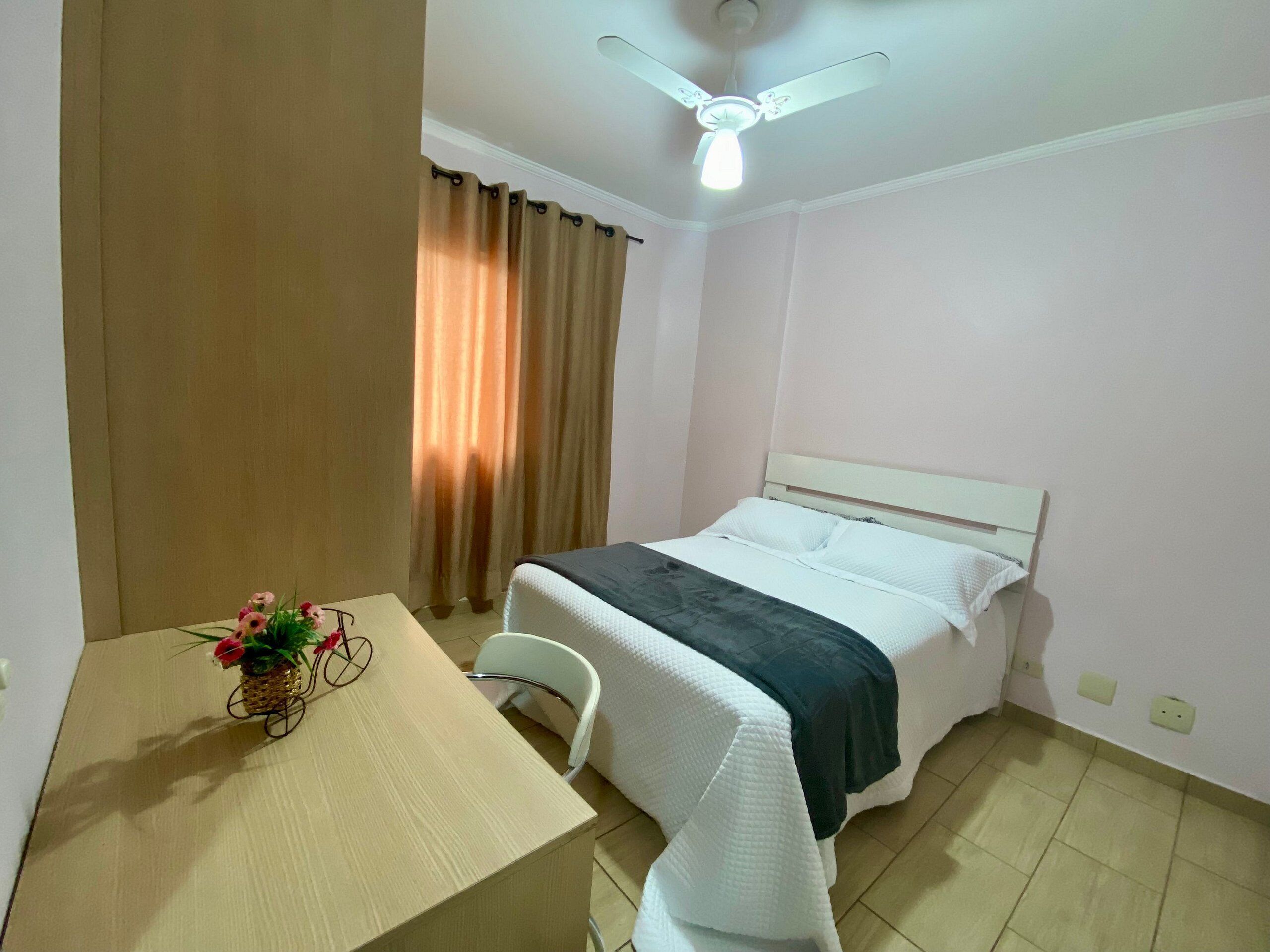 Quarto de hotel com cama, mesa com um vaso de flores, ventilador de teto e cortinas.