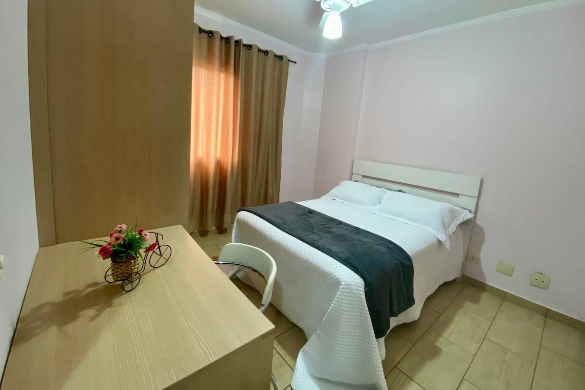 Quarto de hotel com cama, mesa com um vaso de flores, ventilador de teto e cortinas.