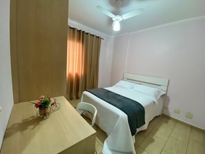 A hotel room with a bed, a table with a vase of flowers, a ceiling fan, and curtains. - Residencial Blaia  (Praia Grande)
