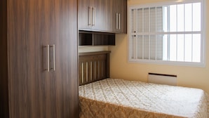 A bedroom with a bed, wooden wardrobe, and a window with blinds. - Residencial Blaia  (Praia Grande)