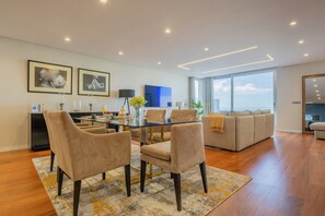 A modern living room with a glass-top dining table, beige sofas, and a large window offering a view of the sky.