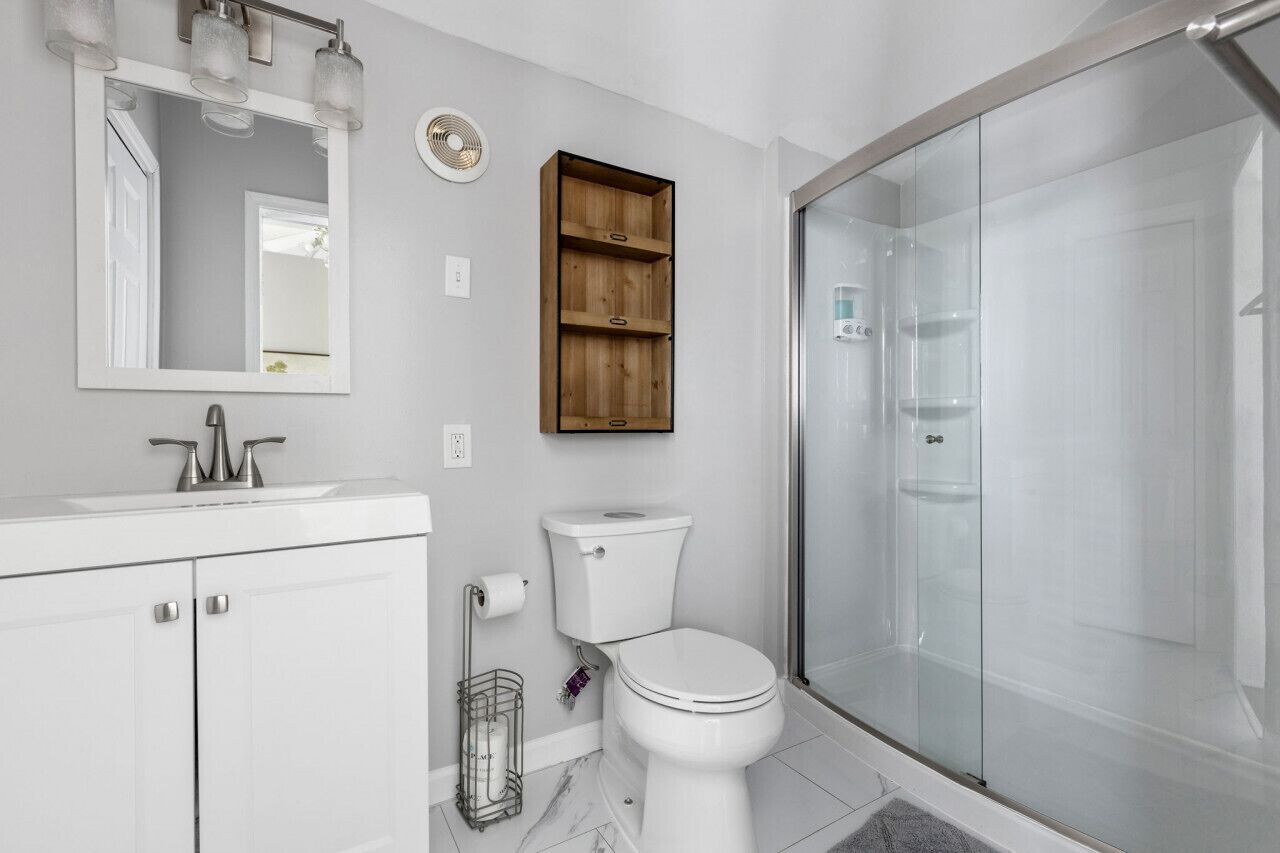 Een moderne badkamer met een wit wastafelmeubel, toilet en een douchecabine van glas. Daarboven bevindt zich een houten plank.