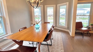 Comedor con mesa de madera, sillas, una araña y ventanas amplias que dan vistas al vecindario.