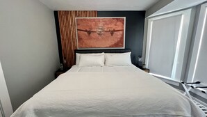 Una cama bien hecha con sábanas blancas, cabecera oscura y un cuadro abstracto grande sobre ella.
