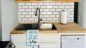 Una cocina con estantes de madera que contienen cuencos color turquesa, platos y un colador. Hay un lavamanos de acero inoxidable con un grifo, un dispensador de jabón para lavar platos y una toalla con un mensaje. Se ve un lavavajillas a la derecha.