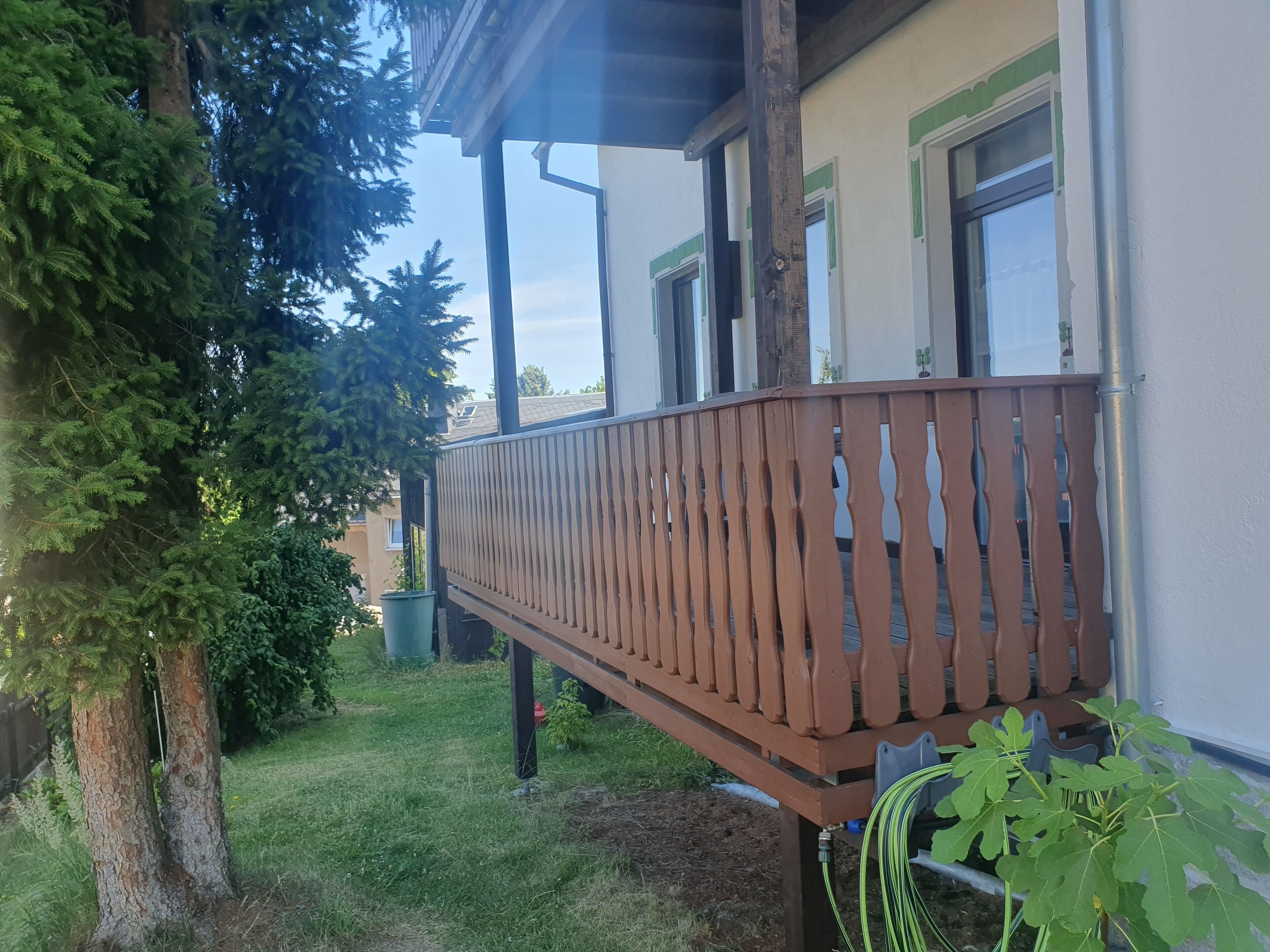 A wooden balcony with a metal railing, a green plant, and a tree in the background.