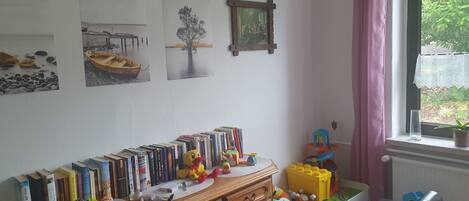 A room with a wooden desk, a window with pink curtains, and various items including books, toys, and a plant.