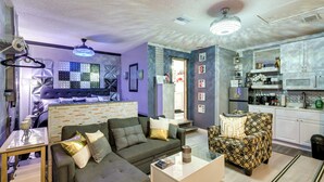 A modern living room with a grey sofa, patterned armchair, and a small coffee table. A kitchenette with white cabinets and a microwave is visible in the background.