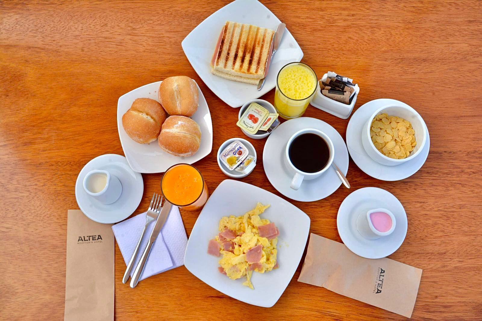 A breakfast setting with bread rolls, a sandwich, scrambled eggs, coffee, juice, and condiments on a wooden table.