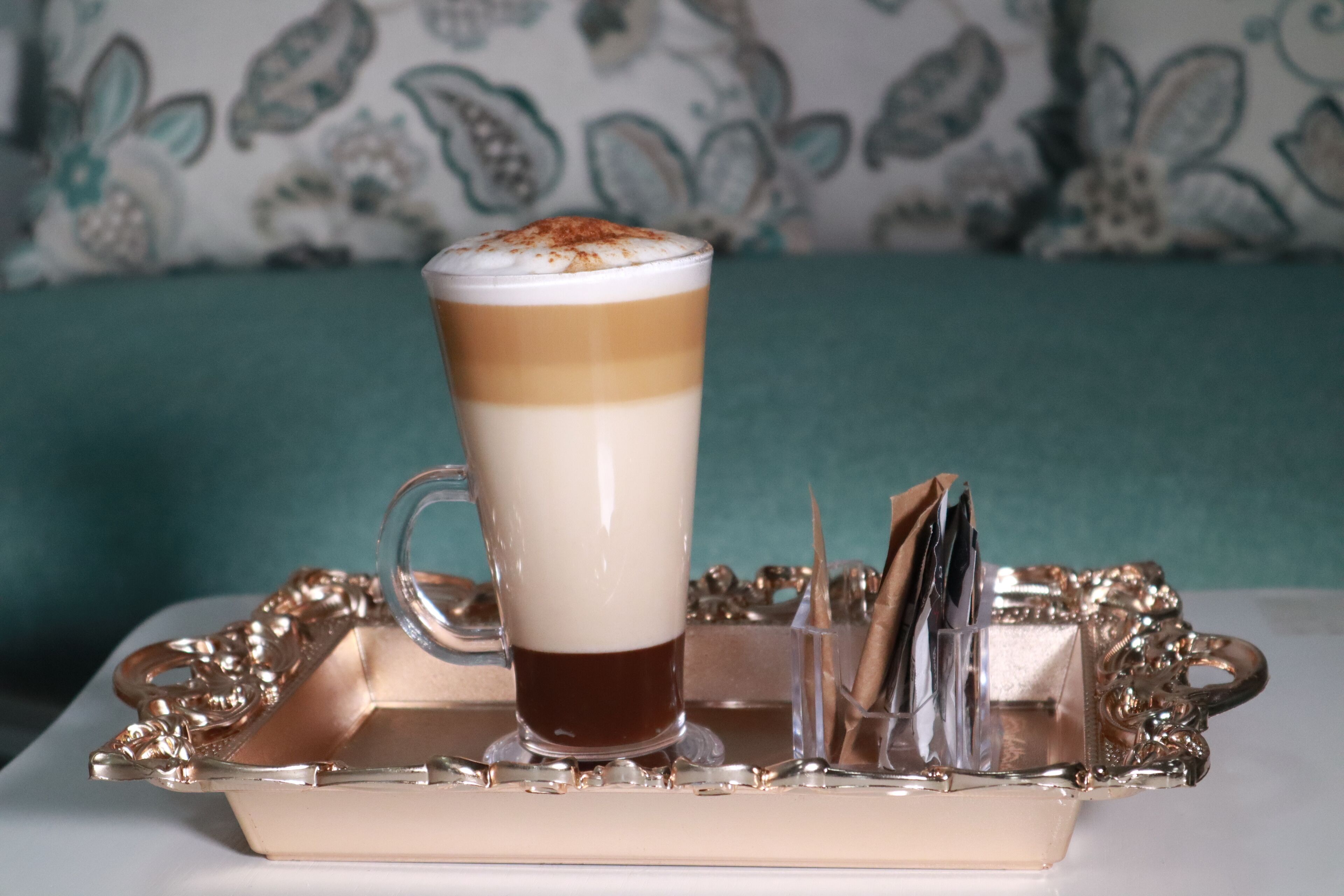 Un vaso alto con una bebida en capas y una espuma en la parte superior, sobre un bandeja decorativa con asa.