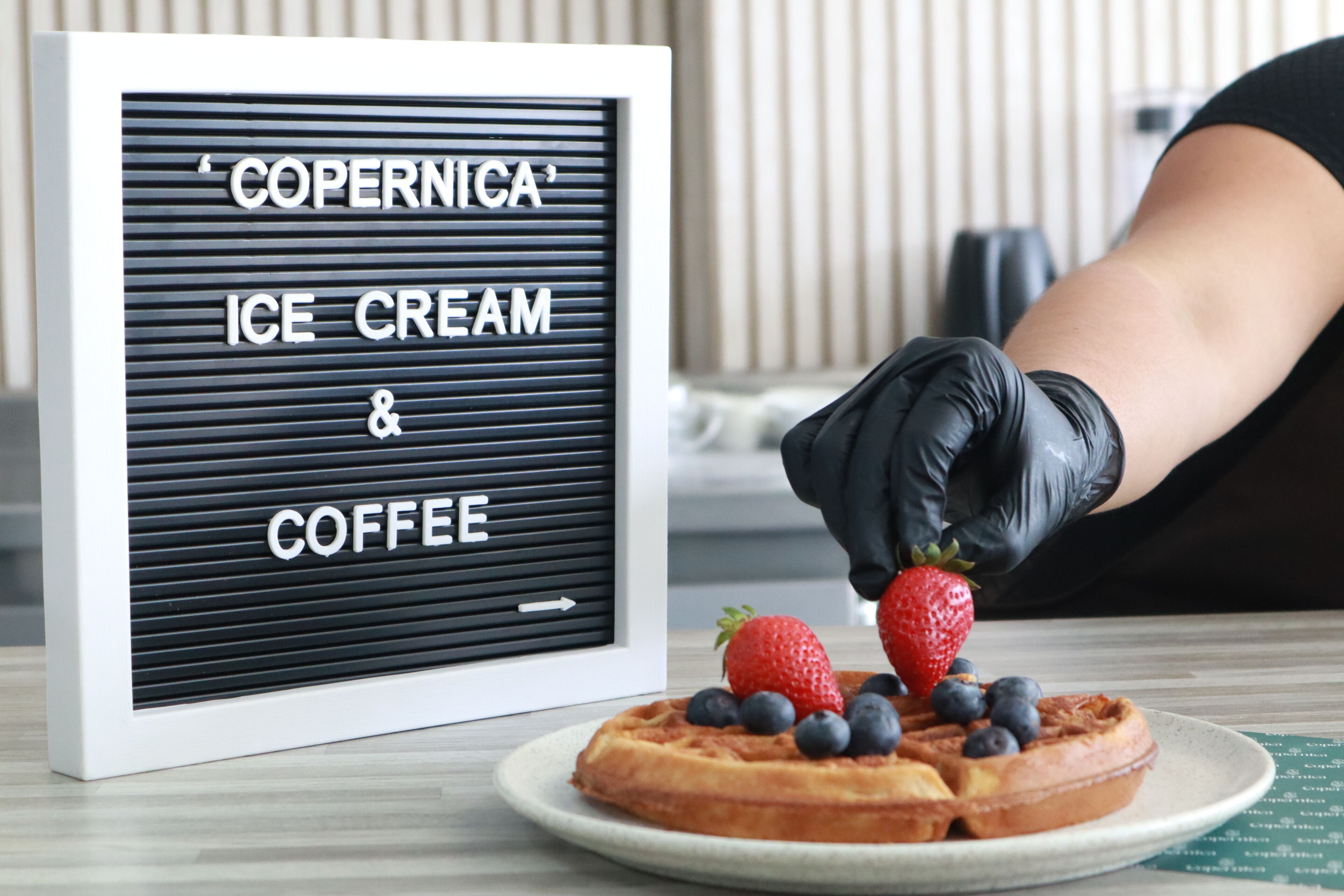 Un letrero enmarcado con la inscripción 'COPERNICA ICE CREAM & COFFEE' y un plato de gofres con fresas y arándanos.