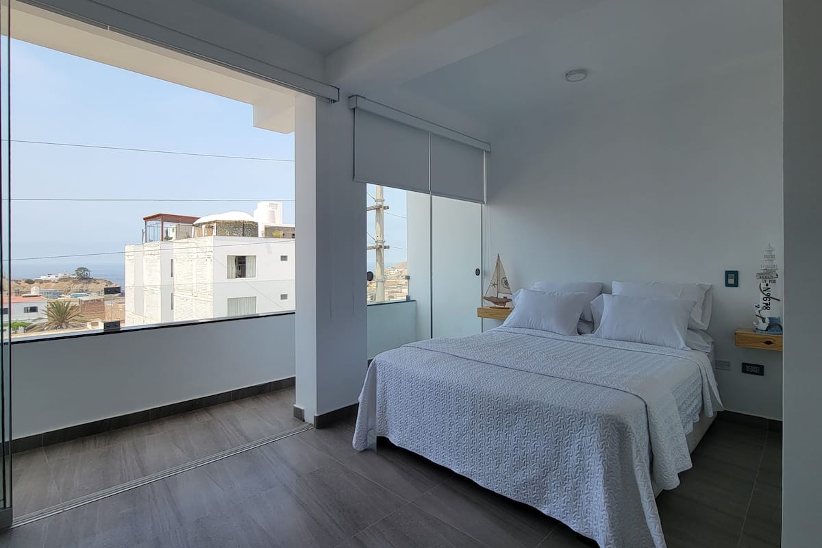 A bedroom with a large window, a bed with white bedding, a nightstand with a lamp, and a view of buildings and the sea.