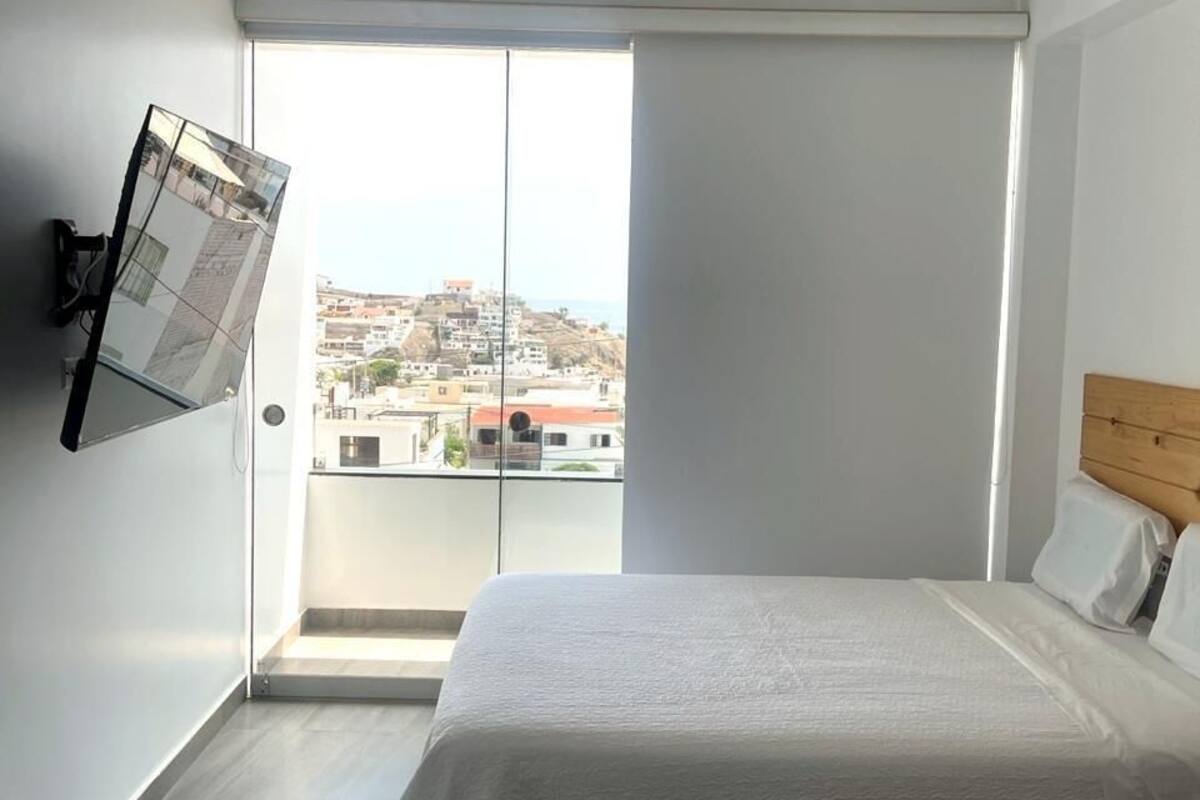 A bedroom with a bed, a TV mounted on the wall, and a view of a cityscape through a sliding door.