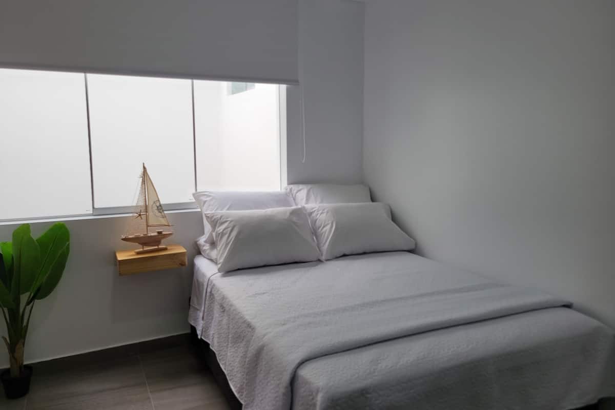 A minimalist bedroom with a bed, a small wooden nightstand, a potted plant, and a window.