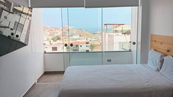A bedroom with a large bed, a view of the city through floor-to-ceiling windows, and a wooden headboard.