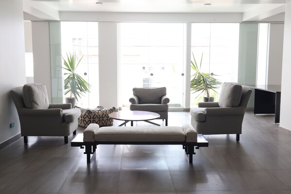 A modern living room with two sofas, a coffee table, and a bench. There are two potted plants and a large window with a view of buildings.