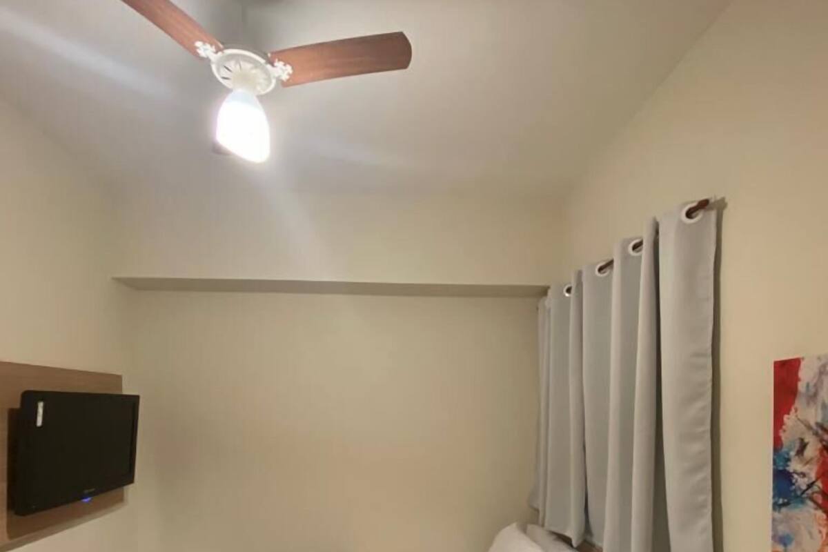 A bedroom with a bed, a ceiling fan, a TV, a small white appliance, and a window with curtains.