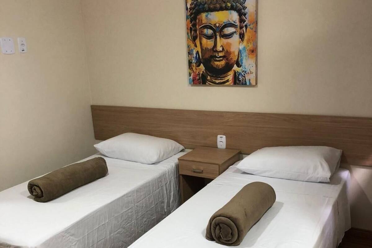A hotel room with two single beds, each with a white bedspread and a brown towel. There is a wooden headboard, a nightstand with a lamp, and a wall-mounted painting of a Buddha.