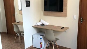 A hotel room with a wooden desk, a television mounted on the wall, a small refrigerator, and a pair of chairs. - POUSADA HD (Nova Friburgo)
