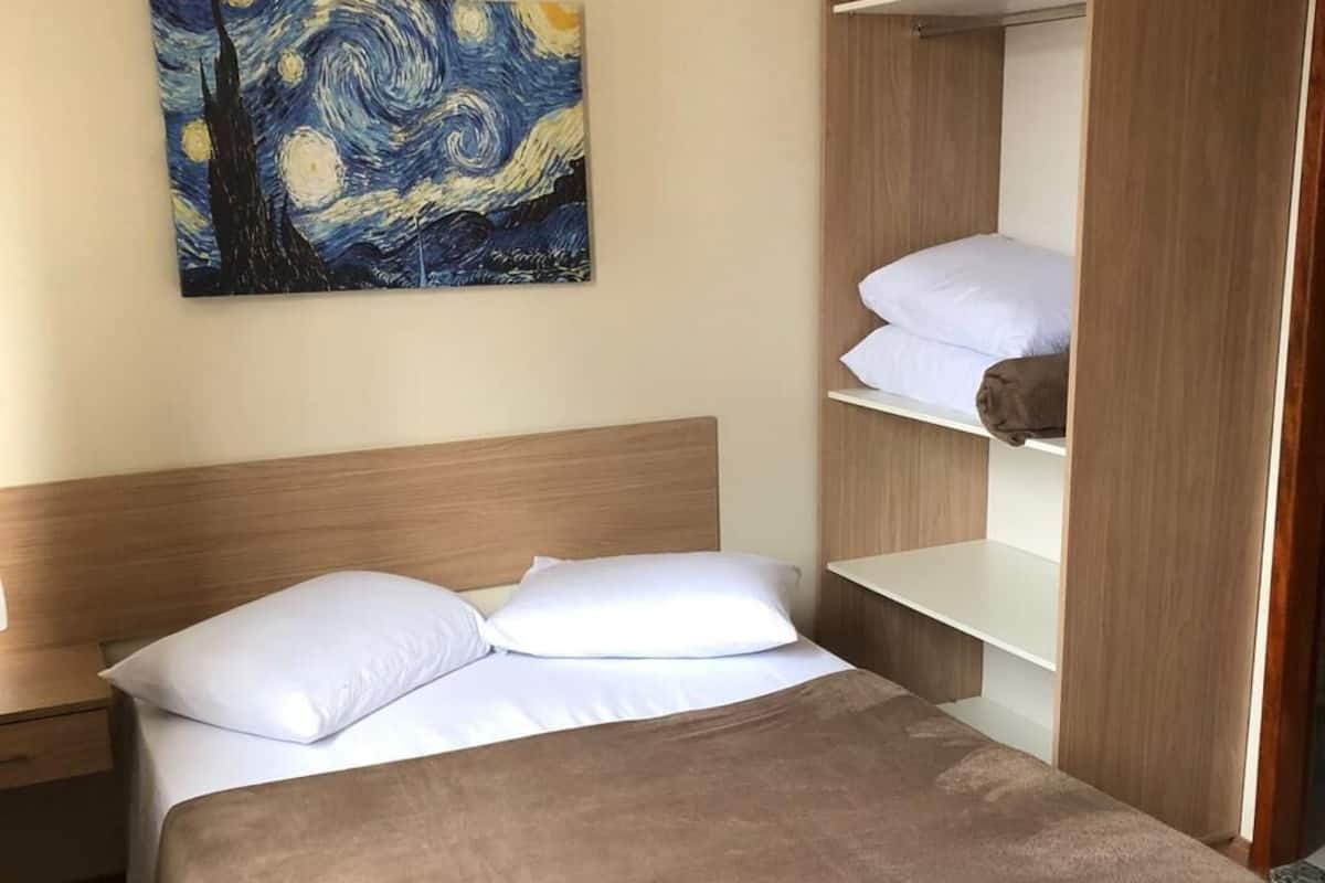 A hotel room with a bed, a nightstand, a painting of a starry night, and a wooden wardrobe with shelves.