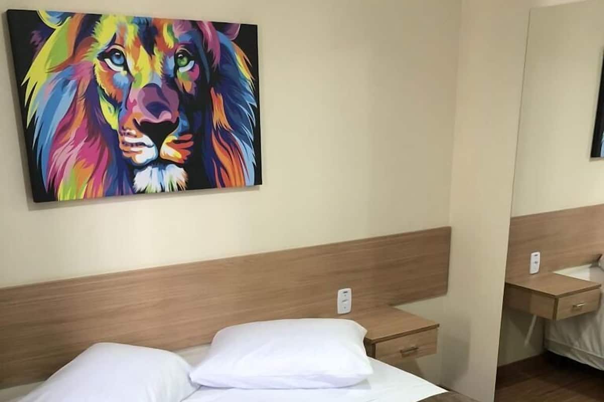 A hotel room with a bed, white pillows, a brown bedspread, a wooden headboard, a bedside table, and a colorful lion painting on the wall.
