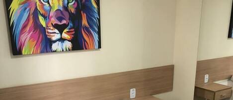 A hotel room with a bed, white pillows, a brown bedspread, a wooden headboard, a bedside table, and a colorful lion painting on the wall.