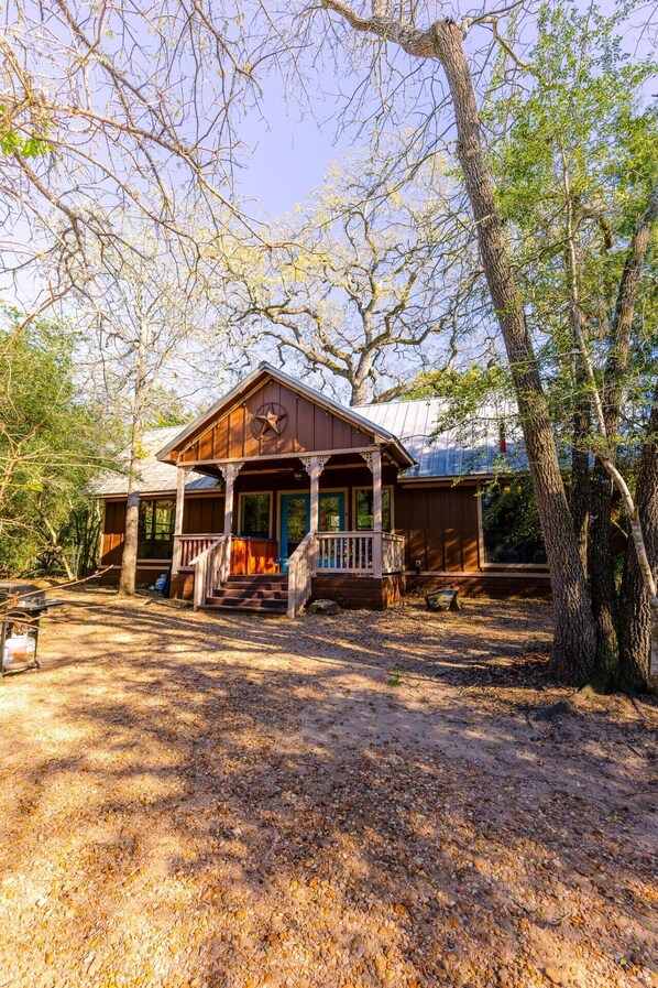 A wooden cabin with a porch, surrounded by trees and a gravel driveway.