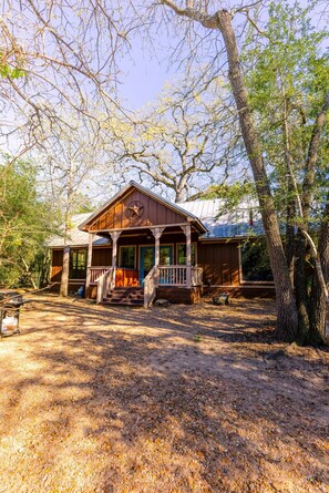 A wooden cabin with a porch, surrounded by trees and a gravel driveway.