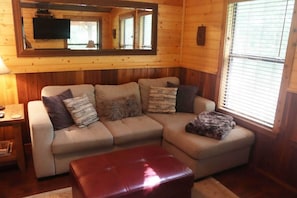 A cozy living room with a sectional sofa, a red ottoman, a TV mounted on the wall, and a window with blinds.