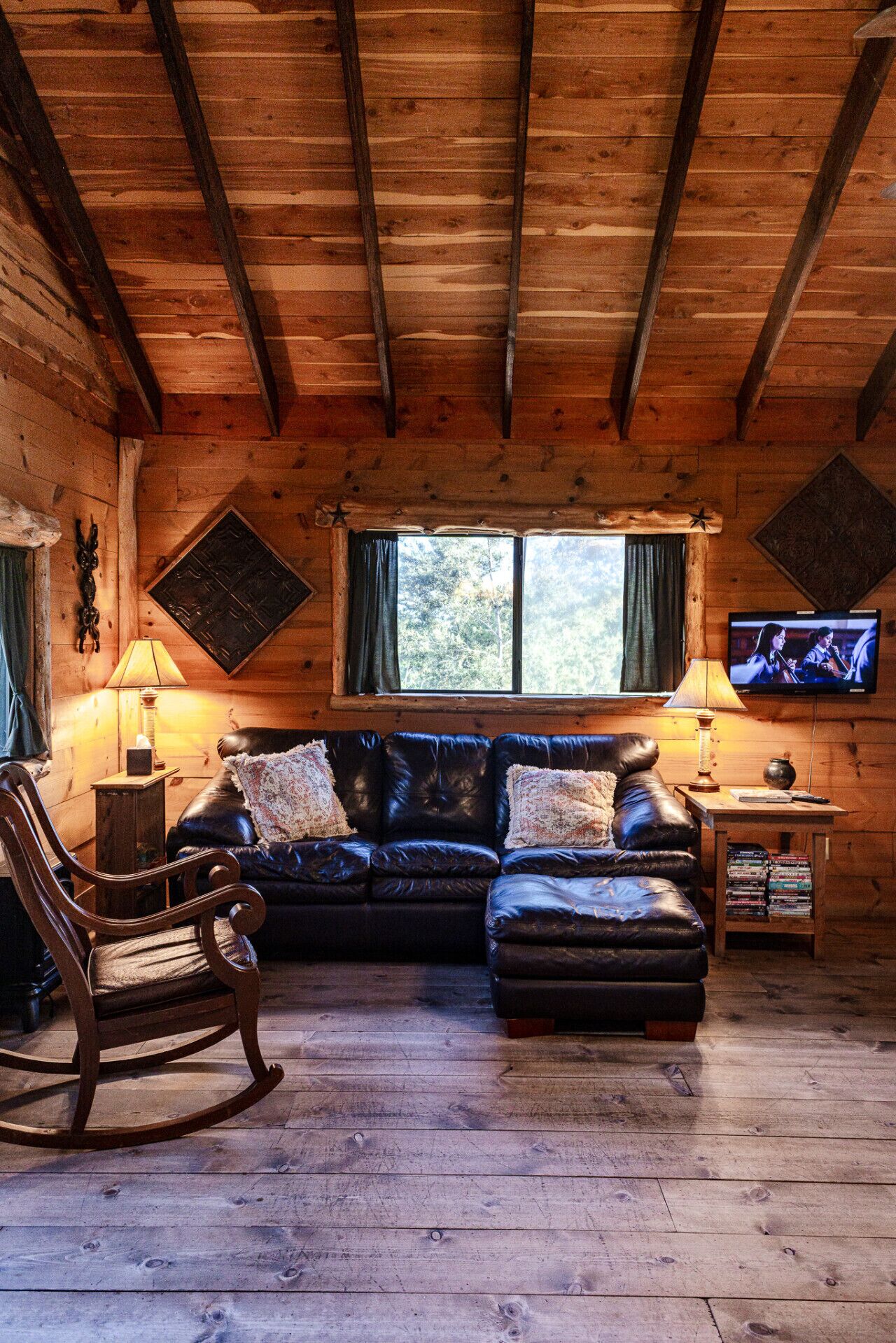 L’intérieur chaleureux d’un chalet avec un plafond en bois, un canapé en cuir, un fauteuil à bascule, un téléviseur et une fenêtre avec des rideaux.