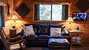 Interior acogedor de una cabaña con techo de madera, un sofá de cuero, un sillón mecedora, un televisor y una ventana con cortinas.