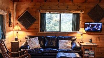 L’intérieur chaleureux d’un chalet avec un plafond en bois, un canapé en cuir, un fauteuil à bascule, un téléviseur et une fenêtre avec des rideaux.