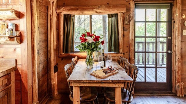 Interior rústico de una cabaña de madera con una mesa de comedor, sillas, un florero con flores y una botella de vino.