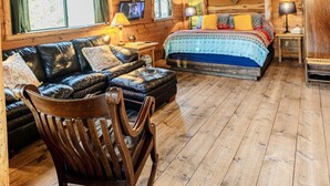 Interior de una cabaña de troncos con un altillo con cama, un sofá de cuero, un sillón mecedor y piso de madera.