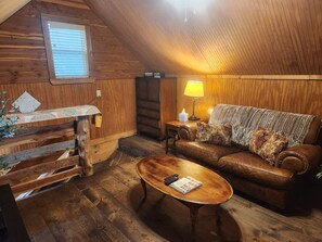 Un accogliente interno di una baita di legno con pavimento in legno, un divano in pelle, un tavolino in legno, una lampada e una finestra con persiane.