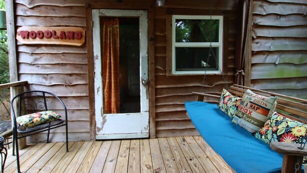 Eine Holzhütte mit einem Schild mit der Aufschrift 'WOODLAND', einer Tür, einem Fenster, einer Bank und einem blauen Sofa mit Kissen.