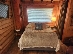 Camera da letto in una baita di legno, con un letto, una finestra con persiane, un comodino con lampada e una sedia.