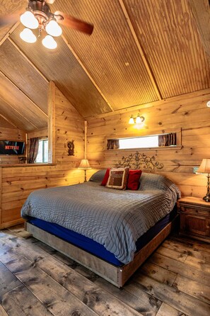 A wooden cabin bedroom with a bed, a ceiling fan, a TV, and a window.