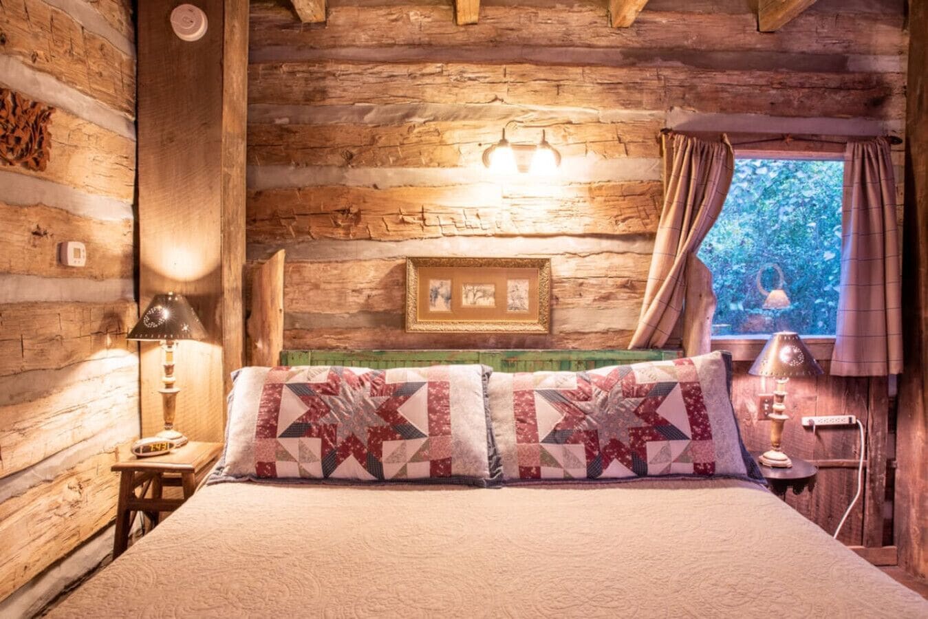 A rustic log cabin bedroom with a bed, two lamps, a framed picture, and a window with curtains.