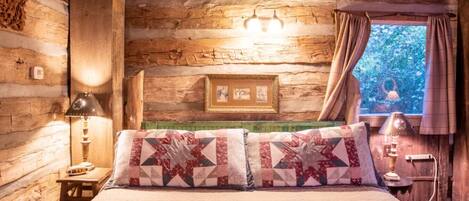 A rustic log cabin bedroom with a bed, two lamps, a framed picture, and a window with curtains.
