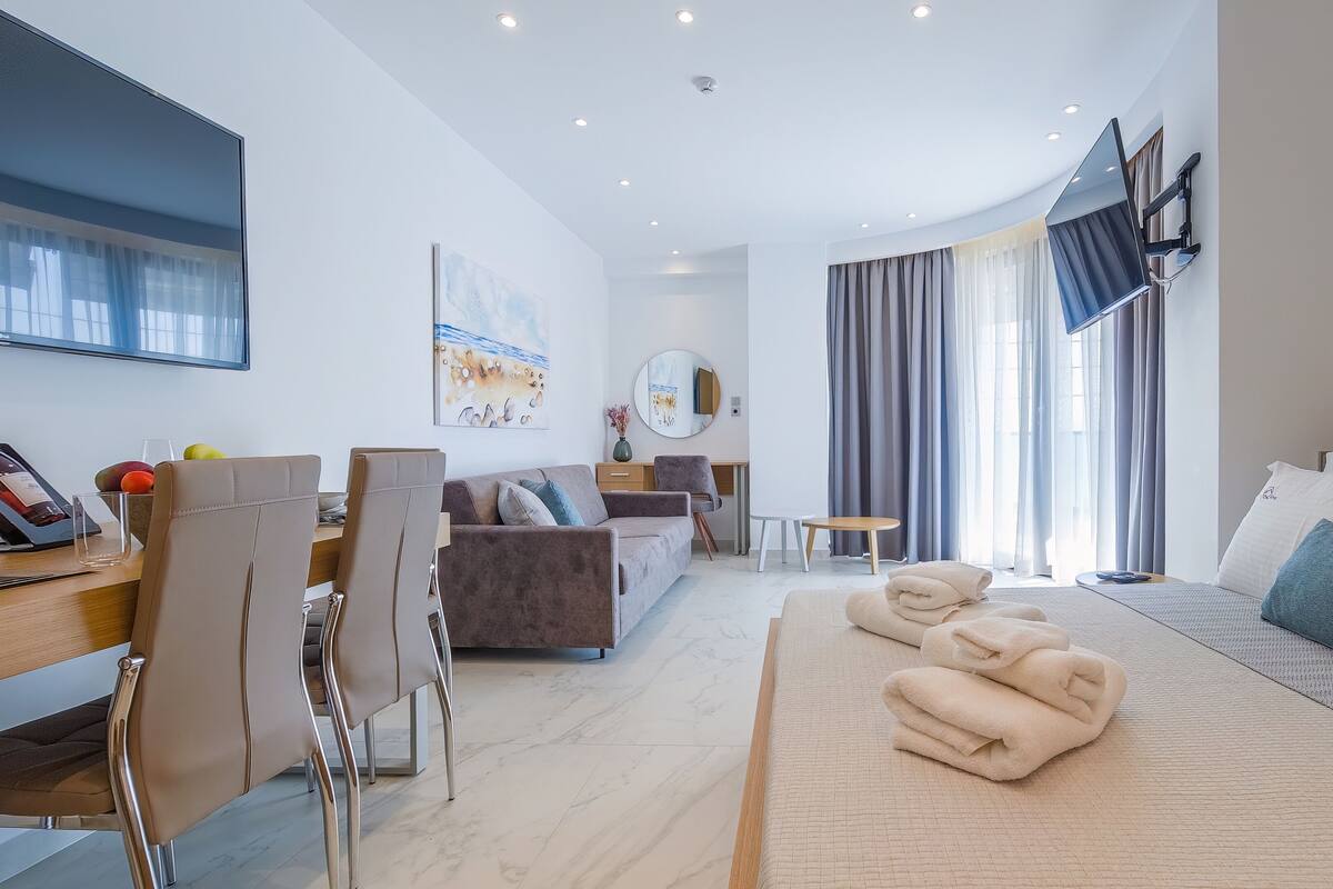 Ein modernes Hotelzimmer mit Bett, Sofa, Essbereich und großem Fenster mit Vorhängen.