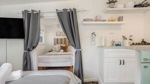 A bedroom with a bed, curtains, a TV, a bedside table, and a small shelf with kitchen items. - Beach Bungalow Parking 2blocks to Beach (San Diego)