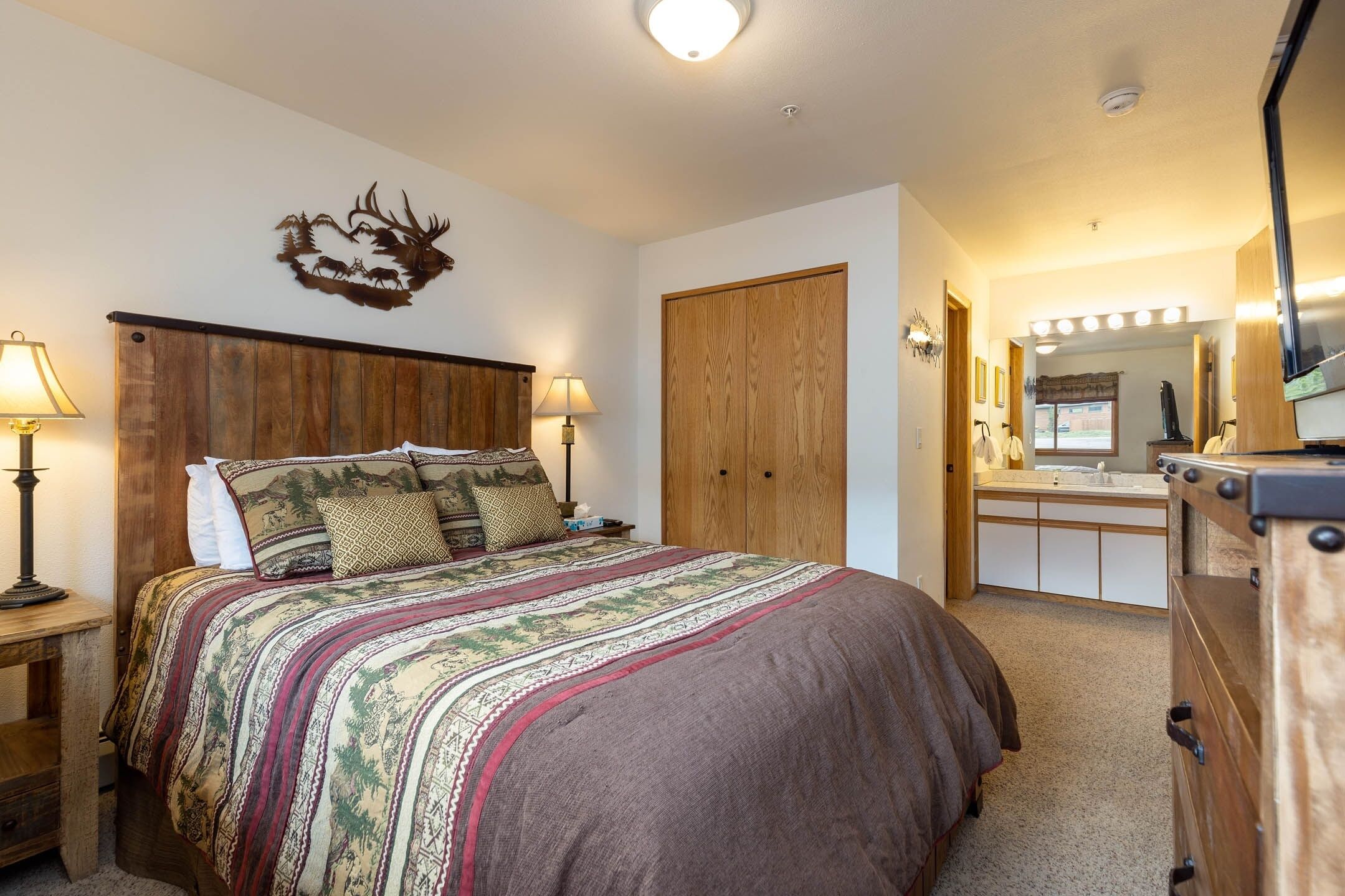 A bedroom with a large bed, wooden headboard, a deer antler wall decoration, and a vanity with a mirror and lights.