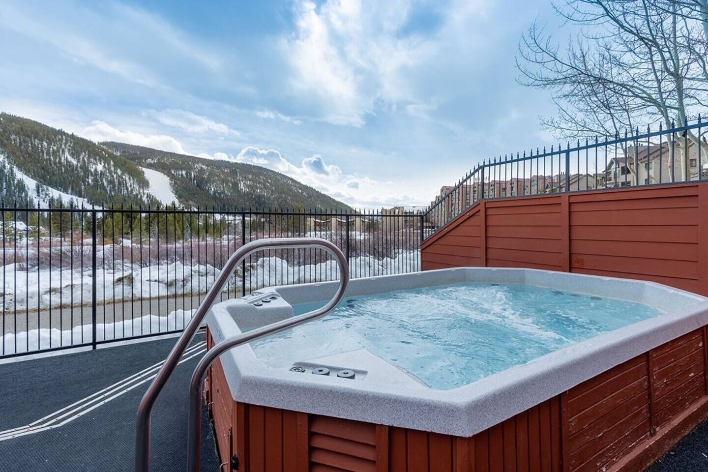 A hot tub with a metal ladder, surrounded by a wooden deck and a metal fence, overlooking a snowy landscape with mountains.